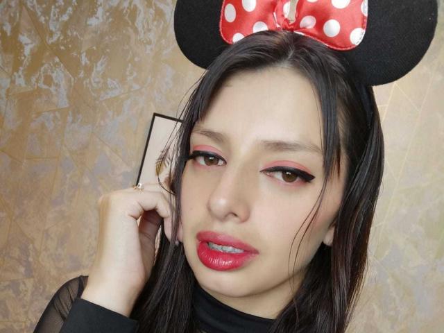 LeticiaFlorenz with black Minnie Mouse ears, red polka dot bow, pink eyeliner, red lipstick, and black top, touching her ear with a hand.