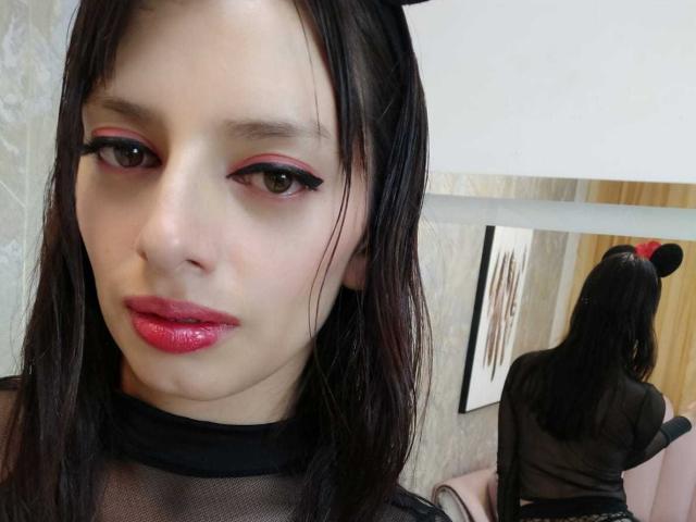 LeticiaFlorenz a la peau pâle, les cheveux noirs et lisses, un fard à paupières rouge, un eye-liner noir, des lèvres rouges brillantes et un haut noir transparent. Ses fesses sont visibles dans le reflet du miroir..