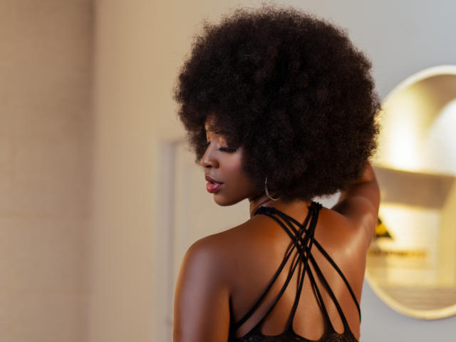 KarmenCastillo has dark, curly afro, wearing a black strappy top, showing her back and looking over her shoulder with a subtle smile, her ass partially visible.