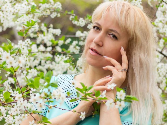 AnaBerry mit blonden Haaren, einem blauen Top mit Punkten und einem Ring, hält weiße Kirschblüten, lächelt sanft, blickt zur Seite, im Hintergrund sind grüne Blätter und Äste zu sehen..