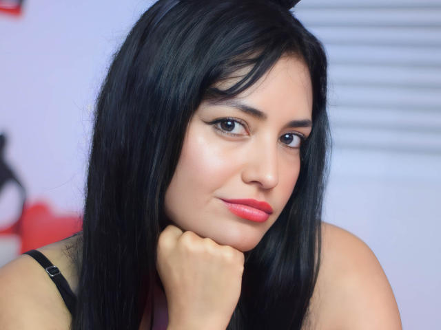 SilvannaReyes has long black hair, red lipstick, and dark eyebrows, with a slight smile, her hand resting on her chin, looking directly at the camera. Her tits are small.