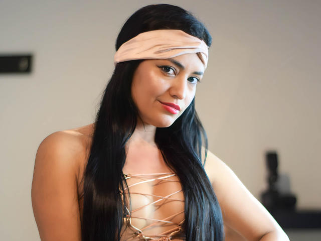 SilvannaReyes heeft lang zwart haar, een roze hoofdband en een gouden top met een ruitpatroon, die haar borsten laat zien, met rode lippenstift en een lichte glimlach, op een foto met zacht licht..