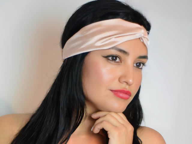 Silvanna Reyes a les cheveux noirs, un foulard rose, et elle regarde sur le côté, sa main près de son menton. Elle porte du rouge à lèvres rose et a un léger sourire..