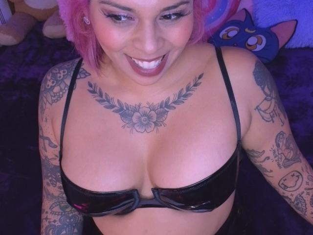 Chili with pink hair is smiling while showing big tits in a black latex bra, displaying a flower tattoo on her chest.