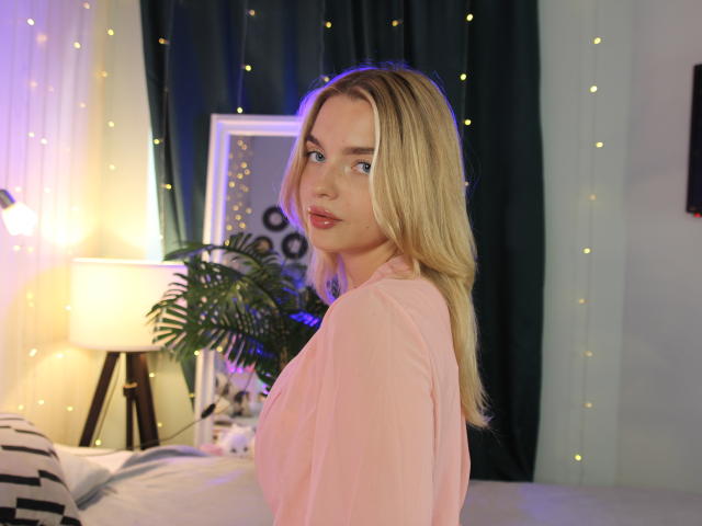 HoneyxRyder mit blonden Haaren, einem pinken Hemd und roten Lippen, die über die Schulter schaut, mit blauen Augen, mittlerer Brustgröße, in einem sanft beleuchteten Raum mit Lichterketten und einer grünen Pflanze..