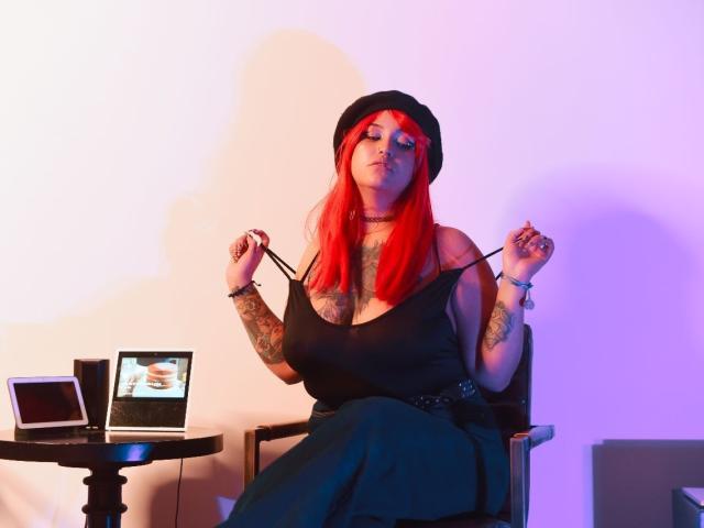 AliBrinck with bright red hair, black hat, black tank top showing big boobs, tattooed arms, pulling straps, laptop on table, sitting, purple and blue light, confident expression.