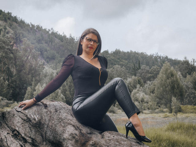 ParisMatur69, avec des cheveux bruns foncés, des lunettes, un haut noir à fermeture éclair, un pantalon en cuir noir et des talons hauts noirs, assise sur un rocher, montrant ses fesses, dans une zone boisée..