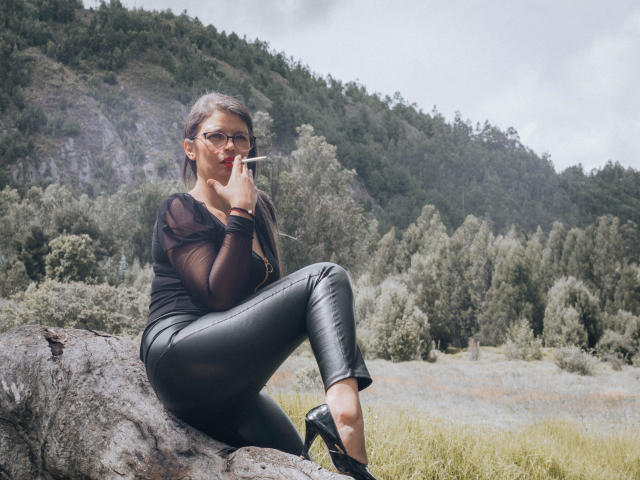 ParisMatur69, con cabello oscuro, gafas, una blusa negra transparente, pantalones de cuero negro y tacones negros, fumando un cigarrillo, sentada sobre una roca, mostrando su trasero, en un bosque..