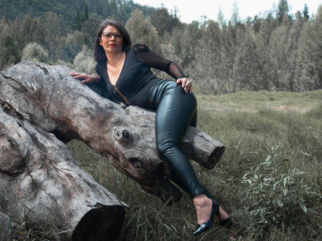 ParisMatur69 con pantalones de cuero negro, una blusa negra, lápiz labial rojo y tacones negros, apoyada en un tronco grande y desgastado en un campo de hierba, usando gafas..