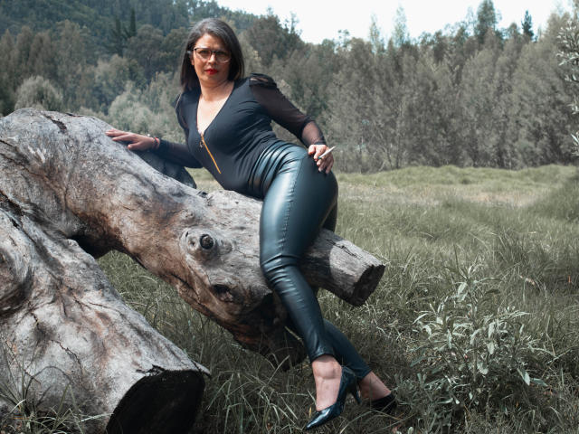ParisMatur69 with red lipstick, black zip-up, and black leather pants, leaning on a large tree trunk, wearing black heels, in a grassy field, forest in background.