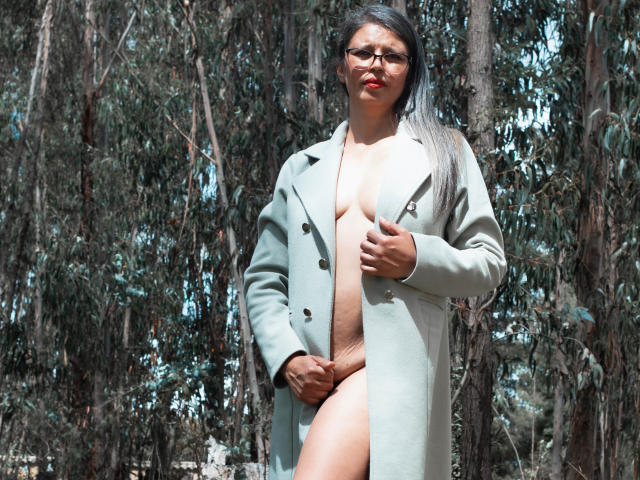 ParisMatur69, avec des lunettes, un manteau gris et du rouge à lèvres, se tient dans une forêt, exposant ses seins nus et ses parties intimes, tenant son manteau ouvert d'une main..
