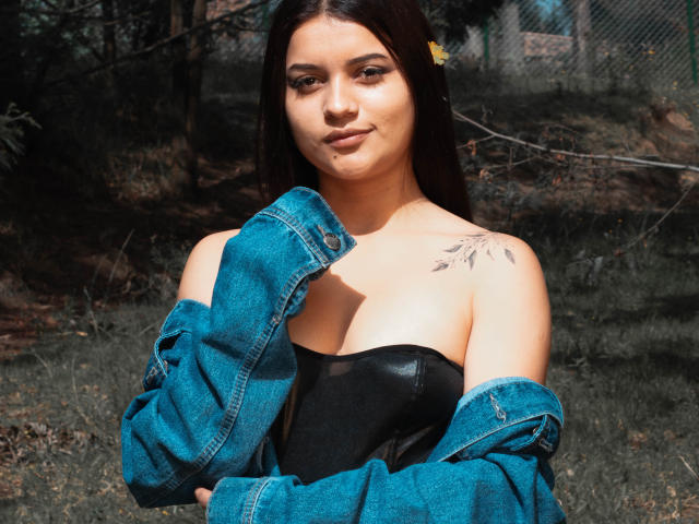 HelenTay, con lunghi capelli scuri, indossa una giacca di jeans blu portata a tracolla sopra un reggiseno di pelle nera, con un tatuaggio sulla spalla destra, in piedi in un'area erbosa illuminata dal sole, sorride leggermente..