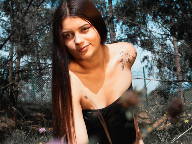 HelenTay, con lunghi capelli castani, un leggero sorriso, un top nero senza spalline, un tatuaggio sulla spalla destra, è in avanti in una foresta, con la luce del sole sul viso, e guarda verso la fotocamera. Il seno è visibile, con luce naturale..