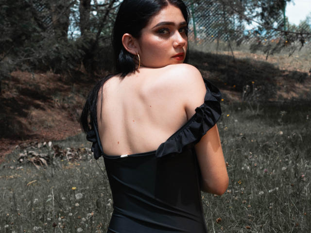 VeroniqueI a les cheveux noirs, le dos nu, et porte une robe noire à une épaule, regardant par-dessus son épaule avec des boucles d'oreilles dorées, dans un décor extérieur verdoyant..