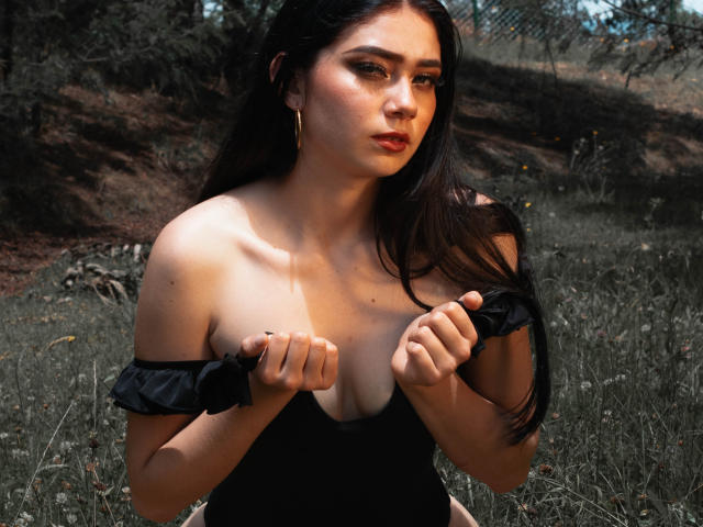 VeroniqueI a de longs cheveux noirs, elle porte un haut noir à une épaule, qu'elle rabat pour montrer ses petites seins. Elle est à l'extérieur, avec de l'herbe et des arbres en arrière-plan..