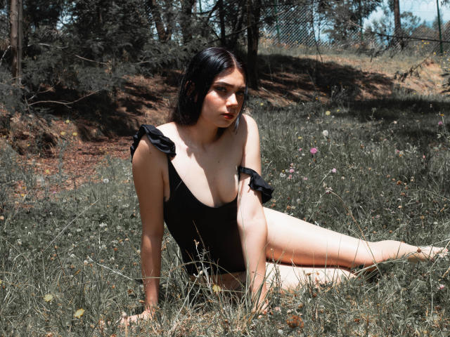 VeroniqueI, con la pelle chiara, i capelli scuri, il seno piccolo e un costume intero nero, è seduta sull'erba, con le gambe distese e le braccia dietro la schiena, e guarda di lato, in una zona boschiva illuminata dal sole..