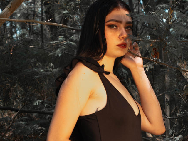 VeroniqueI ha la pelle chiara, i capelli scuri e indossa un top nero con spalline che mette in mostra il suo seno piccolo. Si trova in una foresta, con un'espressione seria e una mano vicino al viso..