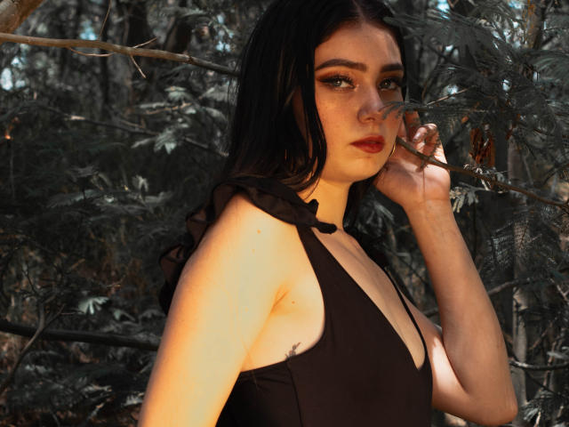 Veronique, avec sa peau pâle, ses cheveux noirs et son rouge à lèvres rouge, portant un haut noir sans manches, a sa main près de son visage, montrant ses seins, dans une forêt..
