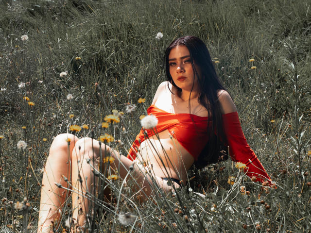 Veronique, con lunghi capelli neri, una maglietta rossa a spalle scoperte e senza pantaloni, seduta in un campo erboso, gambe divaricate, che mostra la sua vulva, circondata da tarassaci gialli e fiori bianchi..
