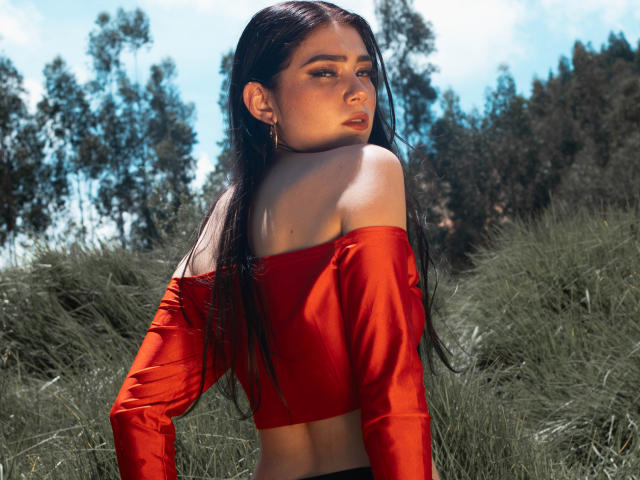 VeroniqueI with long black hair, red off-shoulder top, and gold earrings, looking over her shoulder, showing her bare shoulders and midriff, standing in grassy field.