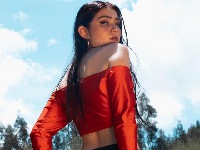 VeroniqueI with long black hair, red off-shoulder top, looking over her shoulder, showing her bare back and upper ass. Bright blue sky and fluffy clouds in the background.