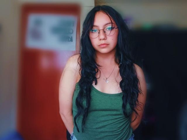 VeroHotLove, avec de longs cheveux noirs ondulés, un débardeur vert, des lunettes, et les mains dans les poches, se tient dans une pièce légèrement floue, et porte un collier avec un petit pendentif..