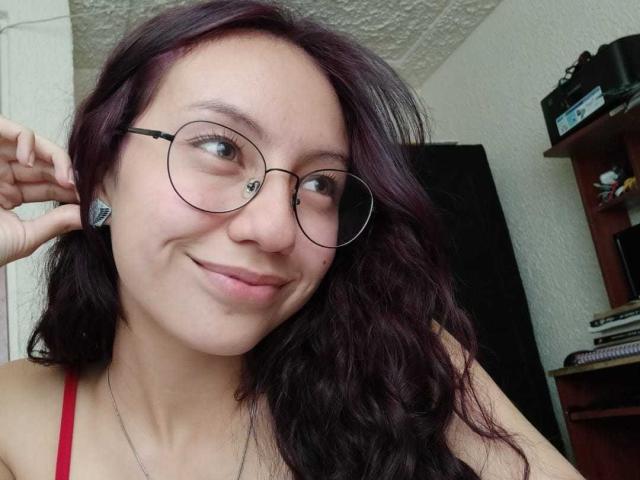 VeroHotLove con capelli scuri viola ondulati, che indossa occhiali rotondi, sorride leggermente, ha una spallina sottile rossa, un orecchino visibile e una collana, e si sta toccando l'orecchio..