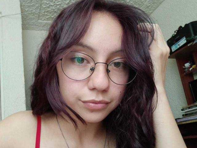 VeroHotLove hat welliges, dunkelviolettes Haar, grüne Augen, runde Brillen und ein rotes Top mit Spaghetti-Trägern. Sie hat eine Hand im Haar und trägt einen neutralen Gesichtsausdruck..