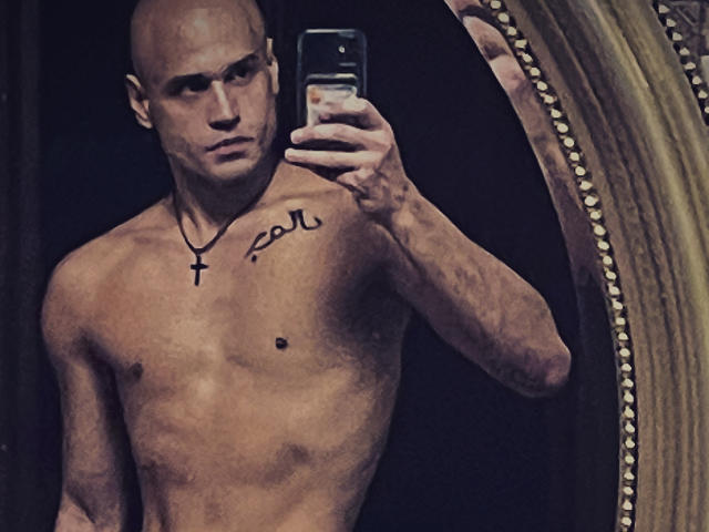 Zamil ohne Hemd, mit glänzendem Kopf und Kreuzanhänger, hält ein Telefon vor einem Spiegel, auf dem ein Tattoo mit kyrillischen Buchstaben auf seiner Brust zu sehen ist..