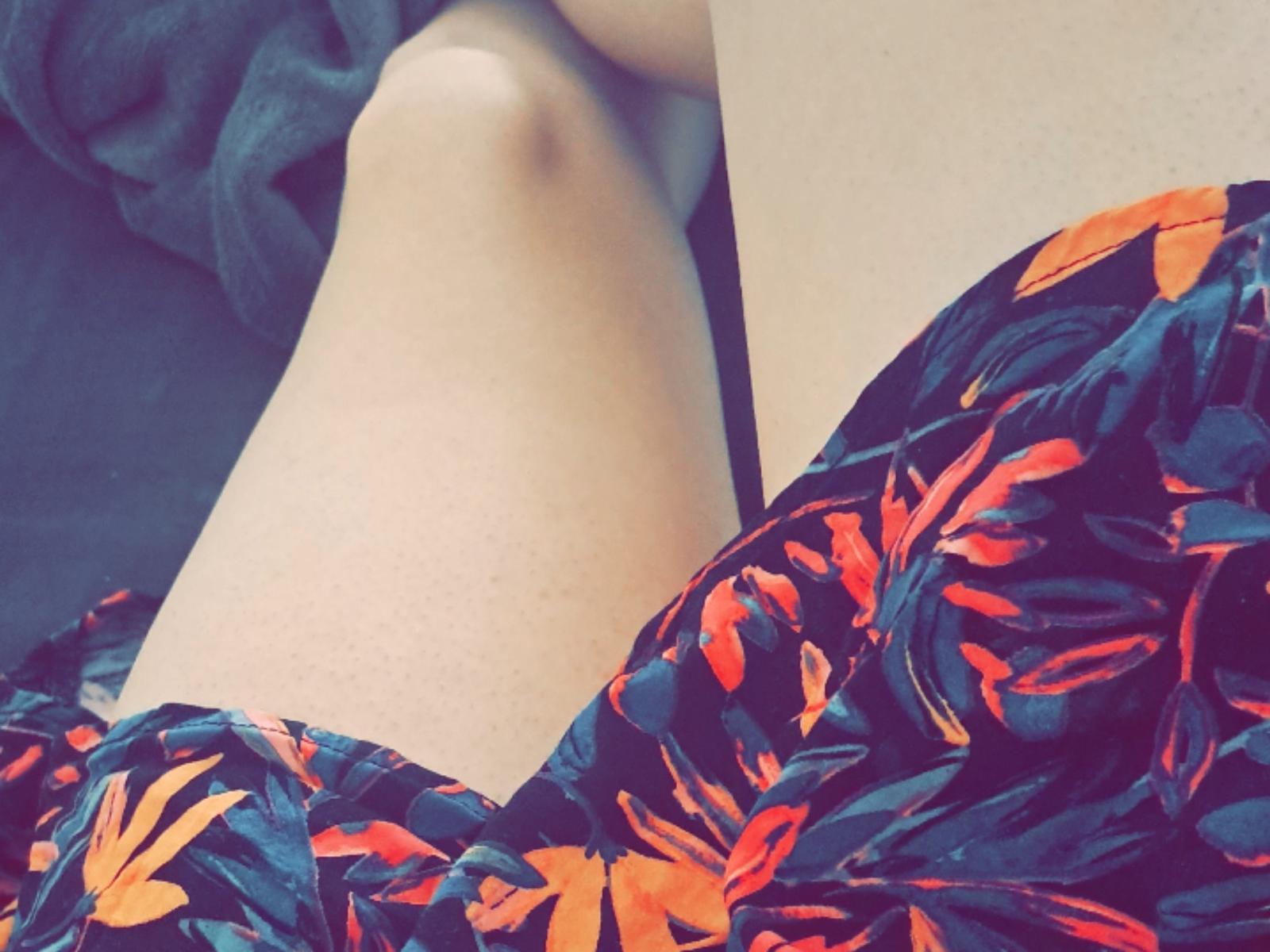 As coxas de NoemieLovesAnal, com pele clara, vestindo um vestido preto com padrões de folhas vermelhas e laranjas, joelho ligeiramente dobrado, tecido macio ao fundo..