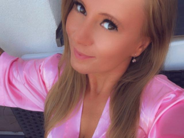 LaMadrina with blonde hair, wearing a shiny pink satin top, smiling at the camera, showing her small tits, wearing pearl earrings, and having light makeup.