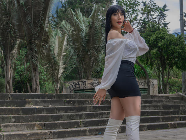 Violeta Parr, avec ses longs cheveux noirs, son haut blanc à épaules dénudées, sa mini-jupe noire et ses bottes blanches montantes, se tient sur des marches en pierre, un téléphone à l'oreille, souriante, et montrant ses fesses..