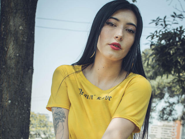 Violeta Parr, con cabello negro largo, camisa amarilla, lápiz labial rojo y aretes de aro dorados, mostrando un tatuaje en el brazo derecho, con una expresión de labios fruncidos, de pie junto a un árbol. Los senos, el trasero y los pezones no son visibles..