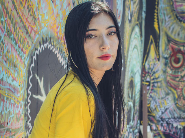 Violeta Parr, con lunghi capelli neri, indossa una camicia gialla, rossetto rosso e orecchini a cerchio, e guarda con occhi scuri. Sullo sfondo, un muro con graffiti. La luce del sole illumina il viso..