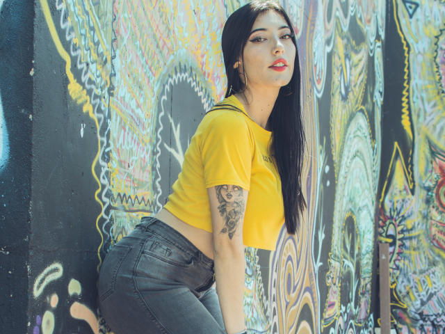 Violeta Parr, con cabello negro largo, un top amarillo y jeans negros, apoyada contra una pared llena de grafitis coloridos, mostrando un tatuaje en su brazo derecho y pintalabios rojo..
