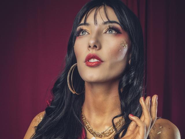 Violeta Parr, met donker, golvend haar, rode lippenstift, gouden oorringen en een gouden ketting, kijkt omhoog met fonkelende ogen en raakt haar wang aan met een glanzende hand..