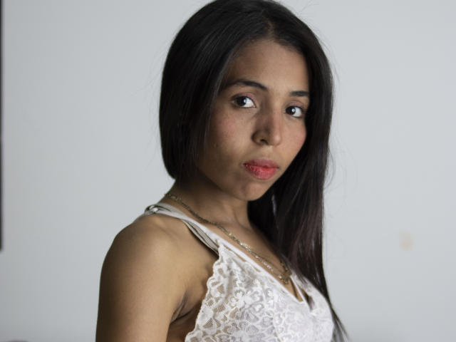 LaylaBrowns with dark brown hair, medium brown skin, wearing a white lace top, looking slightly annoyed with red lips, in a simple photograph.
