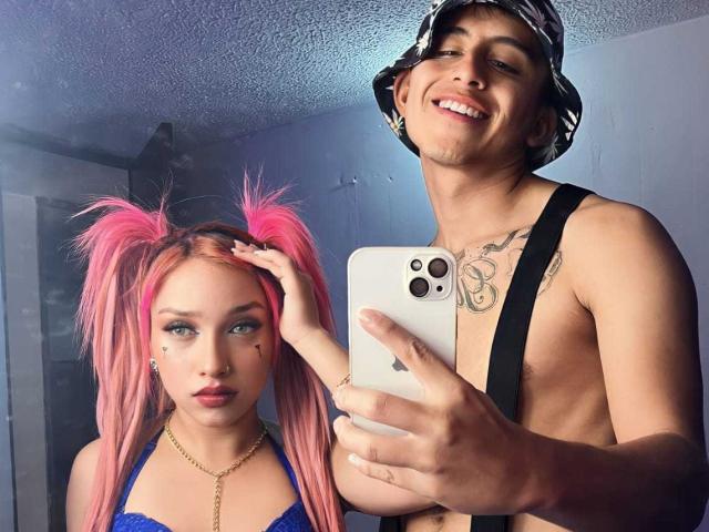 Pinkandkey69, avec ses couettes roses, son haut bleu et son collier doré, se tient à côté d'un homme torse nu portant un seau, des bretelles noires et des tatouages, et qui sourit en prenant un selfie devant un miroir..