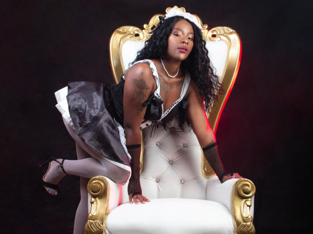 Alanabrant, dark-skinned with long black curly hair, wearing a black and white maid outfit, kneeing on a gold and white throne, showing tattoos, with a seductive gaze.