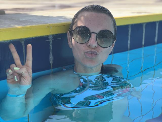 BelleLisaG in a pool with blue water, wearing a floral top, round sunglasses, and puffed lips, flashing a peace sign with a slightly visible pussy through the water.