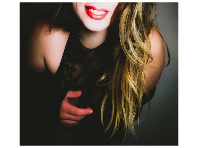 JuliaFrancaise, avec un rouge à lèvres rouge vif, des cheveux blonds ondulés, un haut en dentelle noire, et une main sur ses seins, souriant. Ses seins sont partiellement couverts par la dentelle..