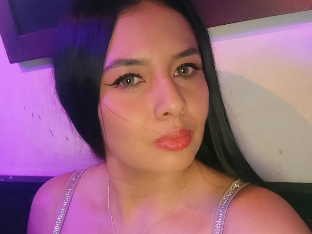 AshleyKatelyn, avec ses longs cheveux noirs, son rouge à lèvres rose et son eye-liner, porte une robe argentée avec des bretelles, et poute ses lèvres pleines, brillantes et roses. Elle a un visage subtil, sexy et mignon..