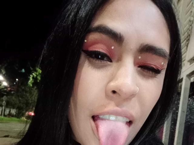 StellaBeckker's face with black hair, pink eyeshadow, nose piercing, and septum ring, winking, sticking out her pink tongue. She has small white studs above her eyebrows.