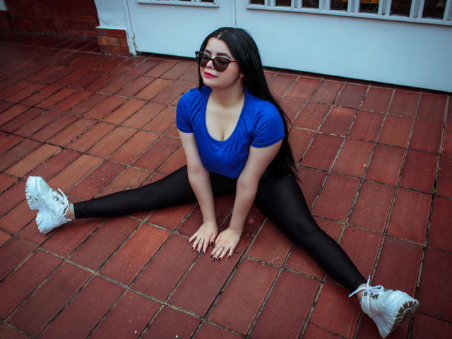 BrithneyLee mit langen, schwarzen Haaren, einem blauen Oberteil, schwarzen Leggings und weißen Sneakers, sitzt auf einem roten Backsteinboden, die Beine weit gespreizt, die Hände auf dem Boden, trägt eine dunkle Sonnenbrille und rote Lippenfarbe..