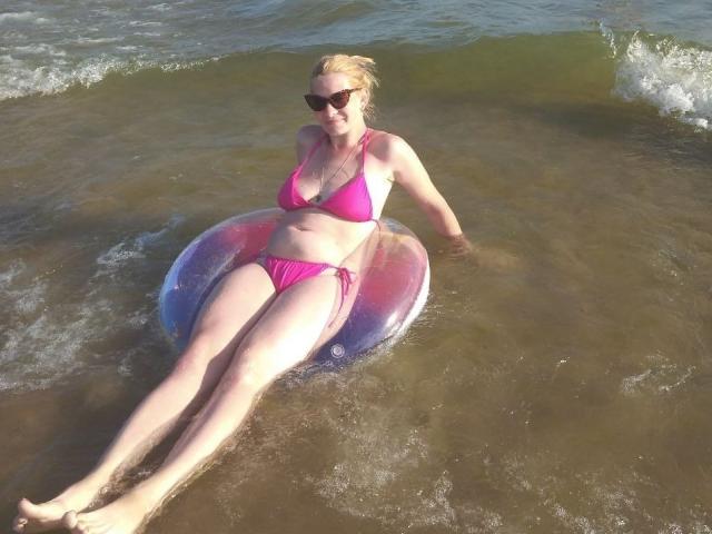 MylesDel mit blonden Haaren, einem pinken Bikini und Sonnenbrille, sitzt auf einem bunten, aufblasbaren Ring im flachen Wasser, die Beine ausgestreckt, leicht feucht, und wirkt entspannt..