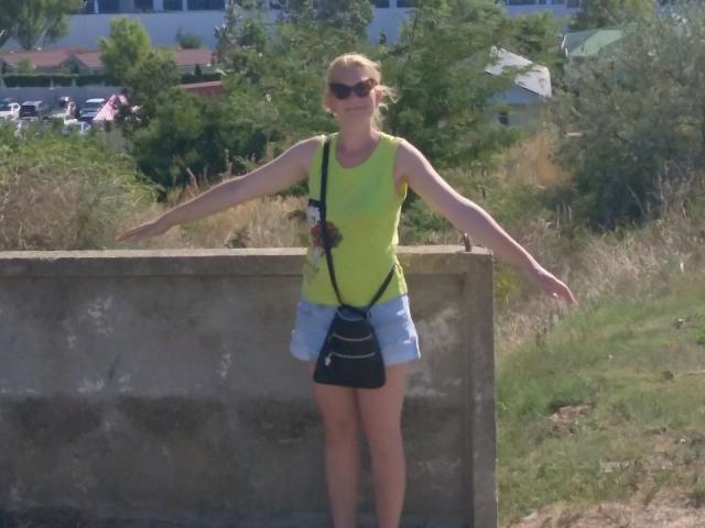 MylesDel, avec des cheveux blonds, des lunettes de soleil, un haut jaune néon, une jupe bleue, un sac à main noir, les bras ouverts, debout contre un mur de béton, à l'extérieur en plein jour..