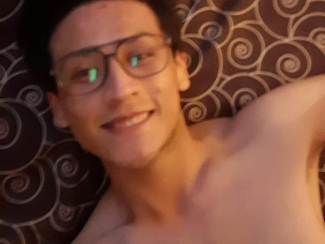 PiesoLores, sin camisa, con gafas, sonriendo a la cámara, reflejo verde en el ojo, pelo corto y oscuro, piel clara, fondo marrón con estampado, clavícula visible, ligeramente borroso, parte superior del cuerpo..