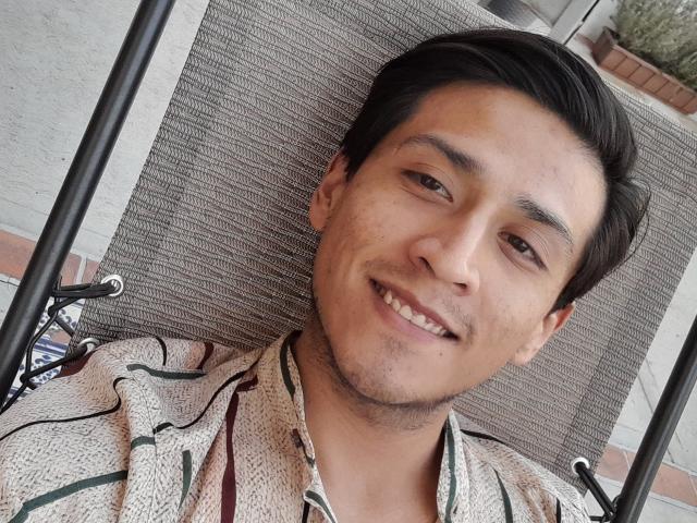 PiesoLores, ein süßer, asiatischer Mann mit kurzen, schwarzen Haaren, lächelt entspannt und trägt ein gestreiftes Hemd. Er sitzt in einem Stuhl mit einer grauen Stoff-Rückenlehne..
