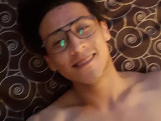 PiesoLores, ohne Hemd, mit Brille, lächelnd, grüne Augen, kurze, dunkle Haare, liegend, gemustertes Hintergrund, weiches Licht, leicht unscharf, lockeres Selfie..