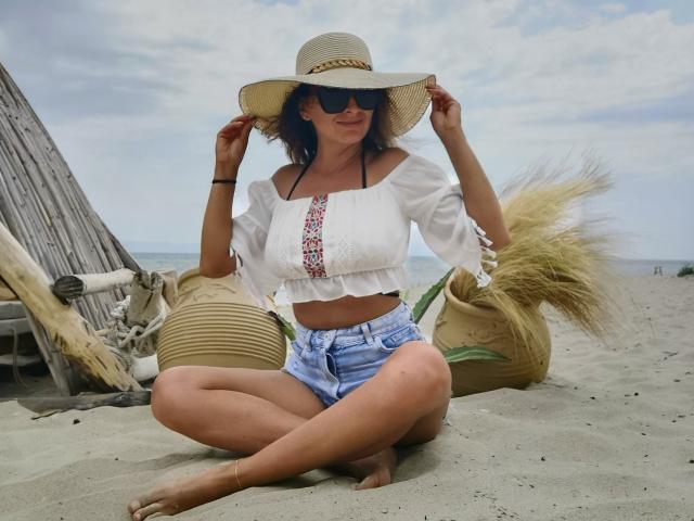 PassionX está sentada en la playa de arena, vestida con una blusa blanca de hombros descubiertos con bordados coloridos, pantalones cortos vaqueros, un sombrero de sol y gafas de sol. Sus manos sostienen el ala del sombrero. Descalza, con las piernas cruzadas..